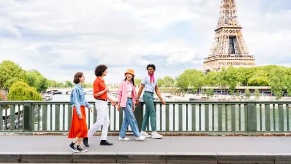 Quatre jeunes amis profitent d'une promenade ensoleillée à Paris, avec la Tour Eiffel en arrière-plan. L'un d'eux, un étudiant, porte un foulard rose vif.