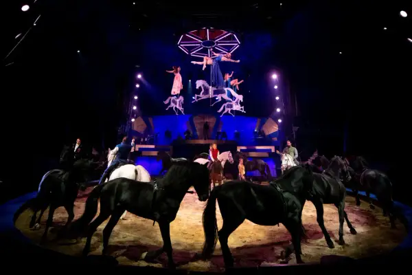 Spectacle équestre des folies Gruss : des chevaux noirs et blancs évoluent sous les projecteurs, tandis que des acrobates aériennes dansent au-dessus.