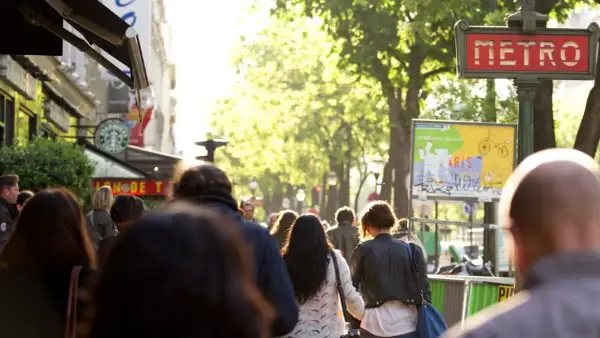Des passants se promènent sur un grand boulevard parisien, sous un soleil radieux. Un panneau "METRO" rouge indique une station.