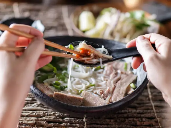 Une cuillère de ce meilleur pho, remplie de nouilles, de viande et d'une touche de sauce piquante, est soulevée avec des baguettes.
