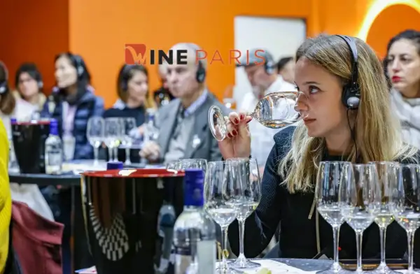 Dégustation de vin à Wine Paris 2026. Une femme hume un verre, casque sur les oreilles, entourée d'autres participants.