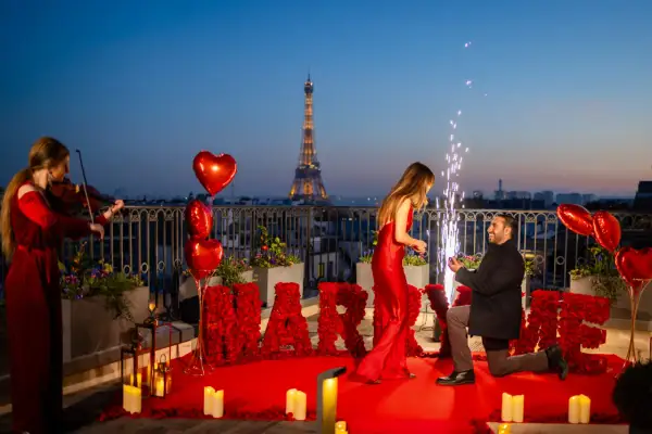 Proposition romantique à Paris, avec la Tour Eiffel en arrière-plan. Un homme s'agenouille, une bague à la main, devant sa compagne. Des roses rouges forment le mot "MARRY ME". Une violoniste joue, des feux d'artifice illuminent la scène. Un moment dig...
