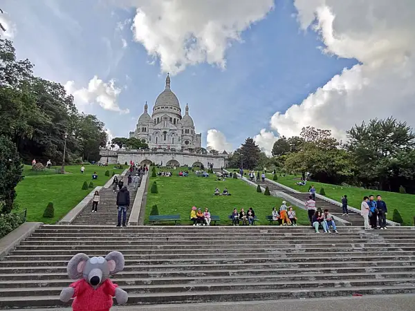 Un adorable raton laveur en pull rouge pose sur les marches menant au Sacré-Cœur, où des visiteurs profitent du soleil sur l'herbe verdoyante.