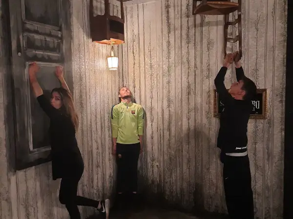 Trois personnes dans une pièce aux murs décrépits, défiant la gravité. Une femme s'accroche à un mur, un homme flotte, un autre escalade une chaise. Ambiance de **the dark dreams Paris**.