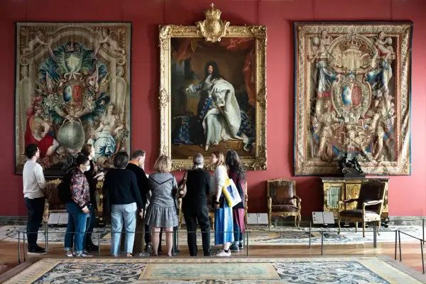 Visiteurs admirant le portrait de Louis XIV et des tapisseries grandioses, une scène digne des 50 plus beaux tableaux du Louvre.