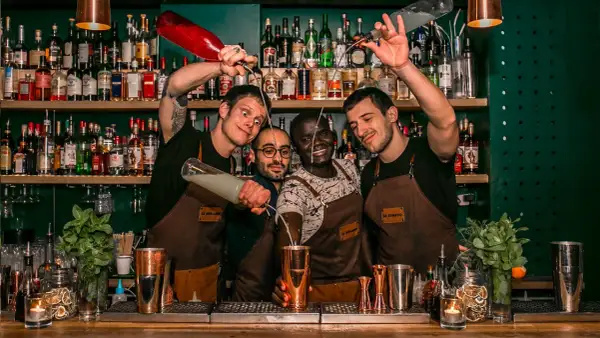 Quatre barmans joyeux préparent des cocktails au bar "Le Renard". L'un verse un liquide d'une bouteille rouge, un autre d'une bouteille transparente.