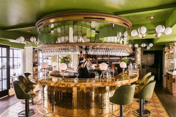 Barman préparant un cocktail dans un bar chic aux murs verts et au comptoir doré. Une ambiance de "pink mama" pour une soirée réussie.