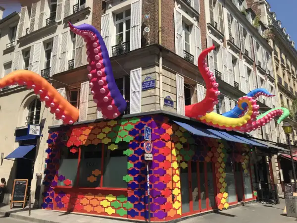 Tentacules colorés géants sortent d'un bâtiment orné de motifs arc-en-ciel, un décor festif pour ce bar gay à Paris.