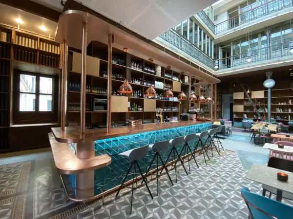 Le bar moderne du "Le Shack Paris" avec son comptoir en cuivre et ses tabourets gris, baigné de lumière naturelle.