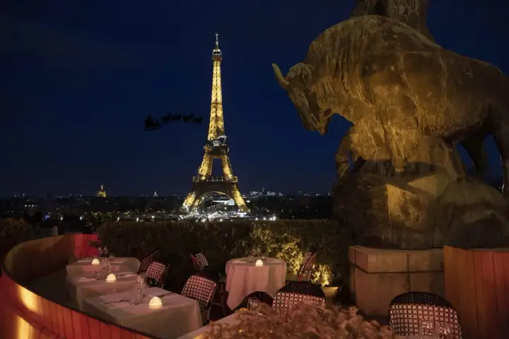 Vue nocturne de Paris avec la Tour Eiffel illuminée. Un bar ouvert le 25 décembre, avec des tables dressées et une statue de taureau. Le traîneau du Père Noël traverse le ciel.